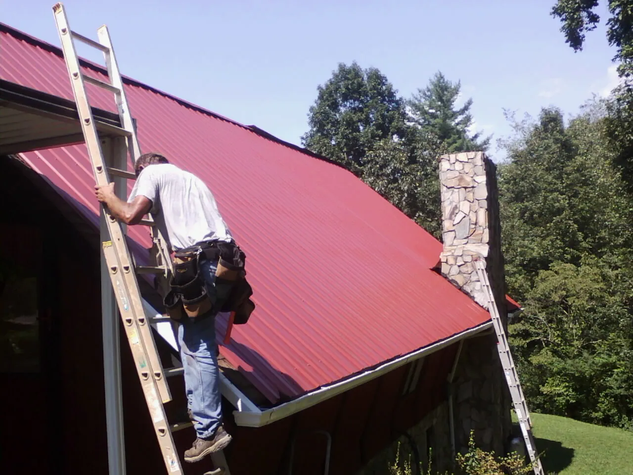 Roofer on a ladder beside a red metal roof and stone chimney — Soffit & Fascia Repair by Roofing Brothers in Port Reading